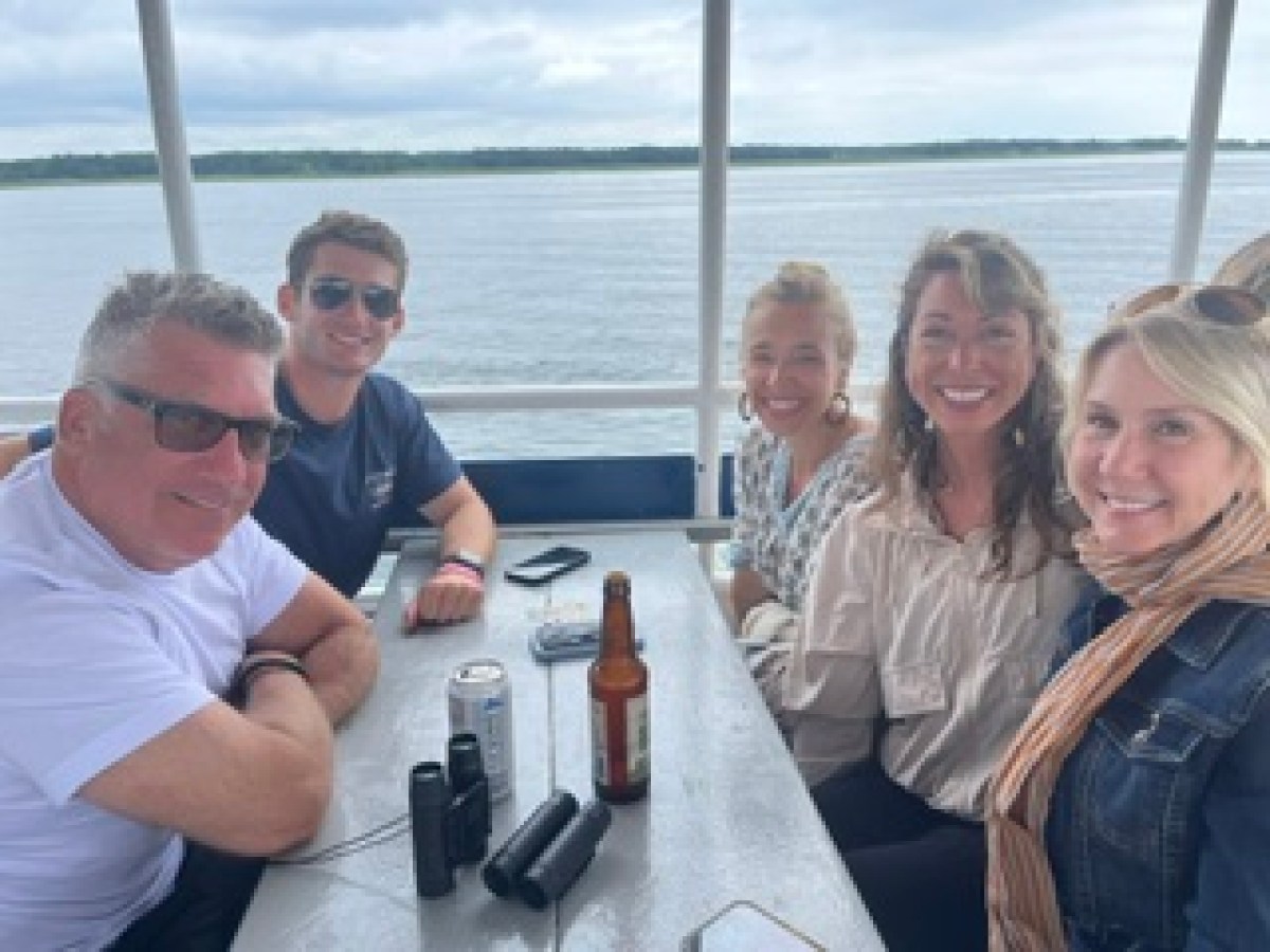 Five people sitting at a table on a boat with water in the background, smiling.