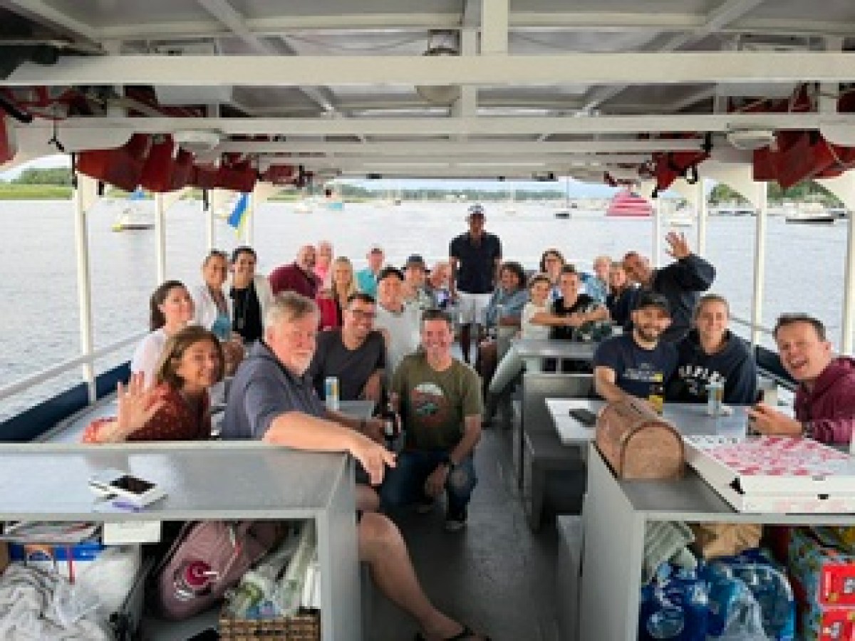 Group of people smiling on a boat with tables, on a calm lake or river.