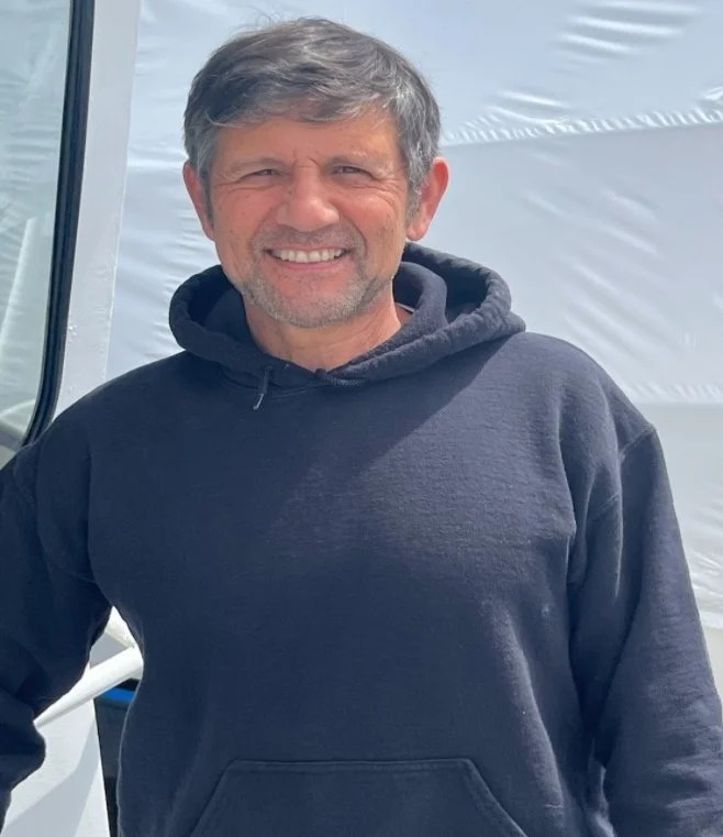 Man smiling in a dark hoodie near a white structure.