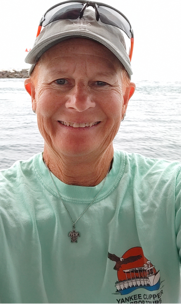 Person in cap and sunglasses smiling near the ocean wearing a nautical-themed shirt.