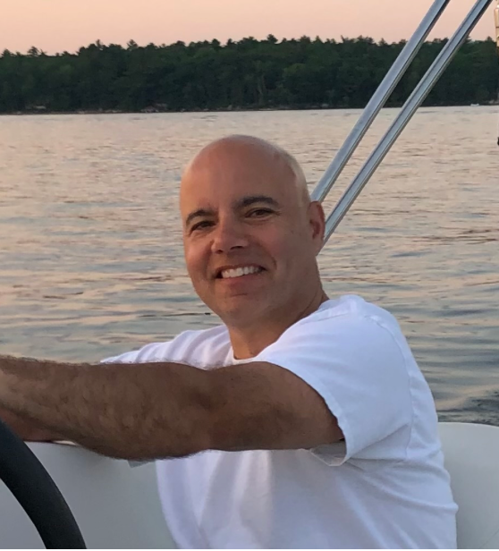 Man smiling on a boat at sunset with trees in the background.