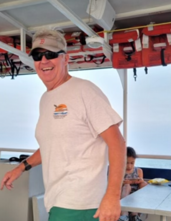 Man in sunglasses and visor smiling on a boat, with a child sitting in the background.