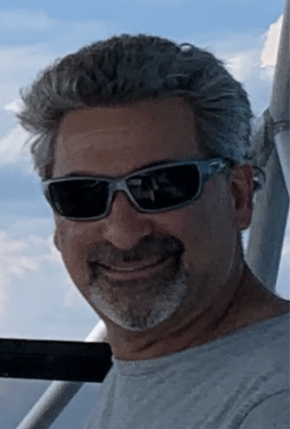 Man with sunglasses and a gray shirt smiling outdoors.