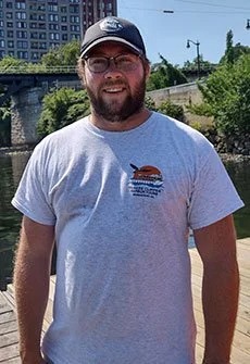 Man wearing a cap and glasses, standing outdoors near water with trees and buildings in the background.