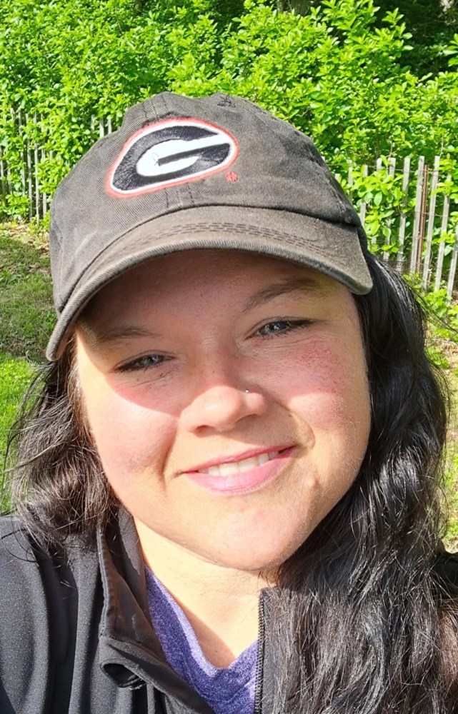 Person with dark hair wearing a black cap and jacket, smiling outdoors.