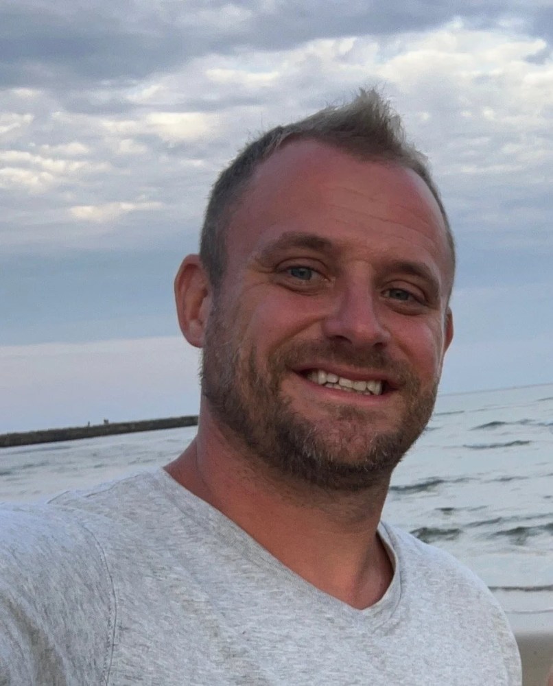 Smiling person with short hair at a beach, cloudy sky background.