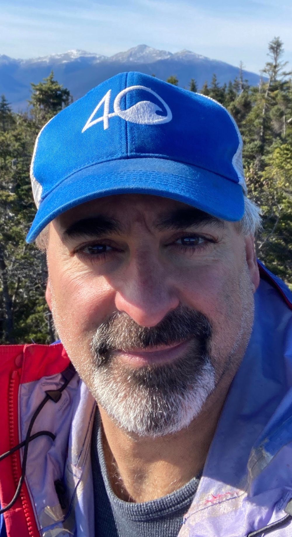 Man in blue cap and jacket outdoors with trees and mountains in background.
