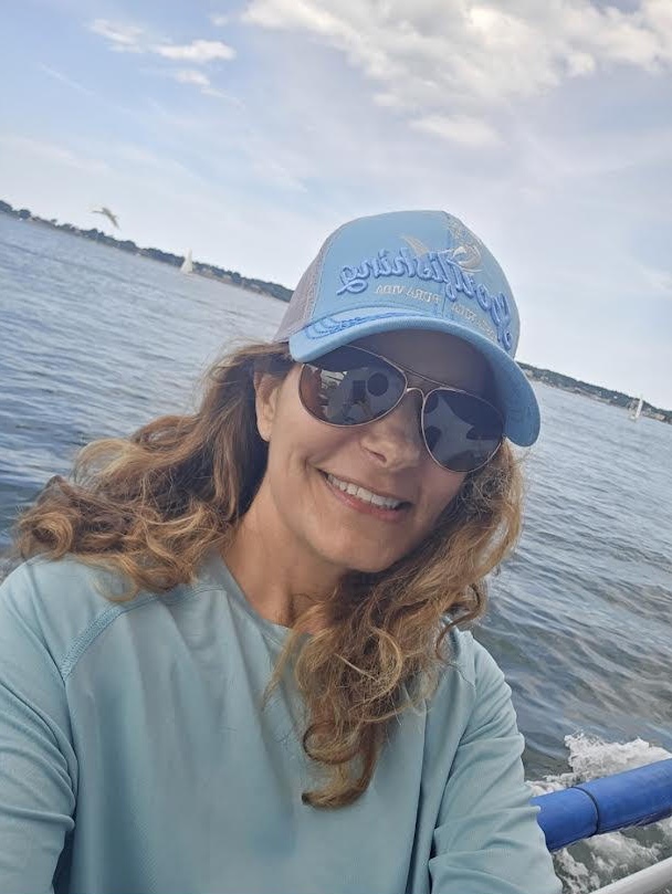 Person in sunglasses and cap smiling on a boat with water and sky background.