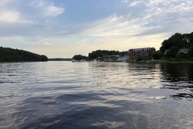 Calm lake with distant buildings and trees under a partly cloudy sky.